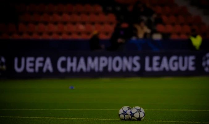 Two soccer balls on a UEFA Champions League field with blurred stands.