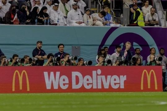 Photographers capturing a sports event behind a McDonald's 'We Deliver' sign.