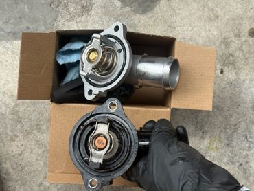 Comparison of a new and old automotive thermostat held over a cardboard box.