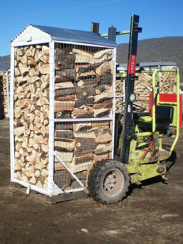 A forklift carrying a large rack filled with stacked firewood outdoors.
