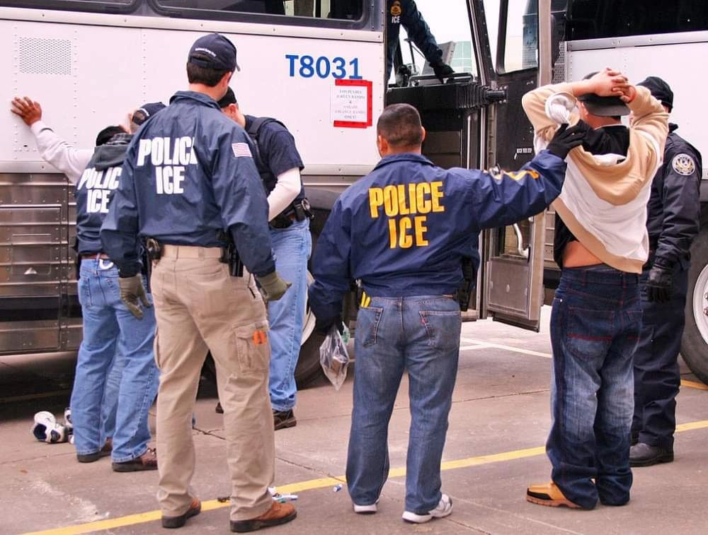 ICE officers detain and search individuals near a transport bus.