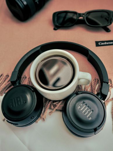 A cup of coffee placed inside JBL headphones with sunglasses nearby.