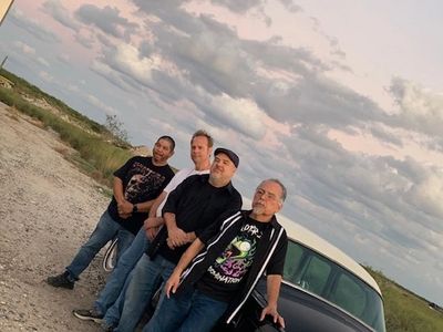 Four men standing beside a vintage car under a cloudy sky.