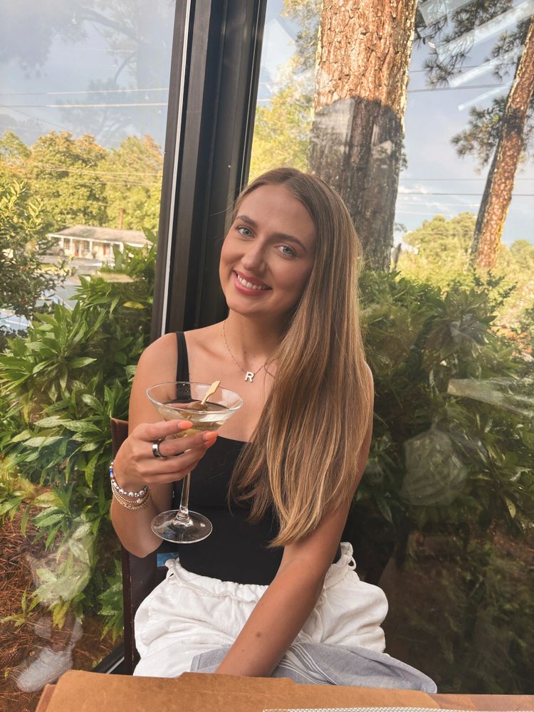 A smiling woman holding a cocktail glass, sitting by a window with greenery outside.