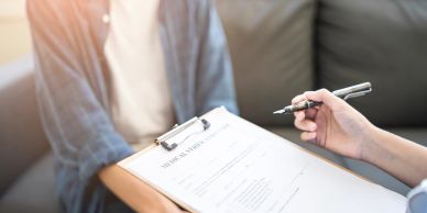 Person filling out a medical form with another person nearby.