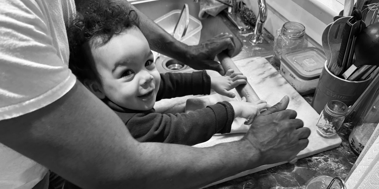 A child joyfully helps an adult roll dough in the kitchen.