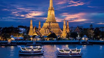 Illuminated Wat Arun temple at dusk with boats on the river in Bangkok.