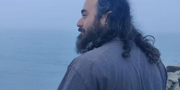 Man with long hair and beard stands by the foggy seaside.
