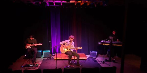 Three musicians performing on a dimly lit stage with purple lighting.