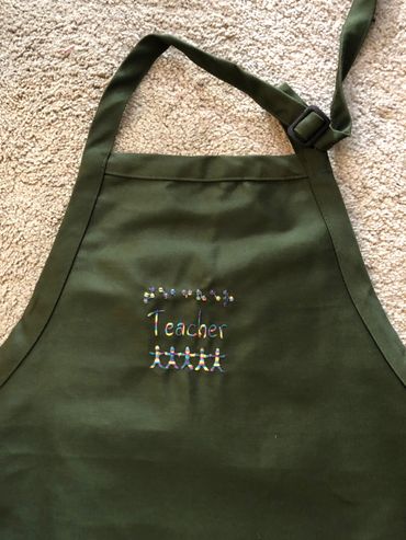 Dark green apron with pockets and adjustable neck. Teacher in Braille and print. Image of children o