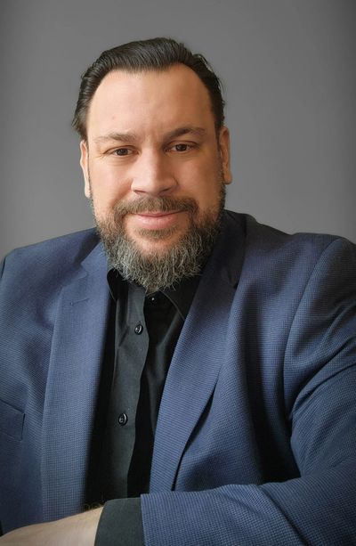 Man in a navy blazer and black shirt smiling warmly against a gray background.