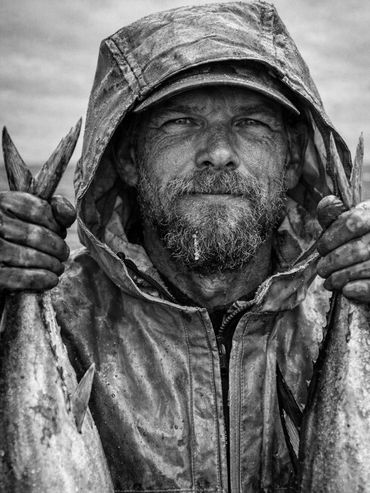 Grizzled fisherman in rain gear proudly holding two fresh fish.