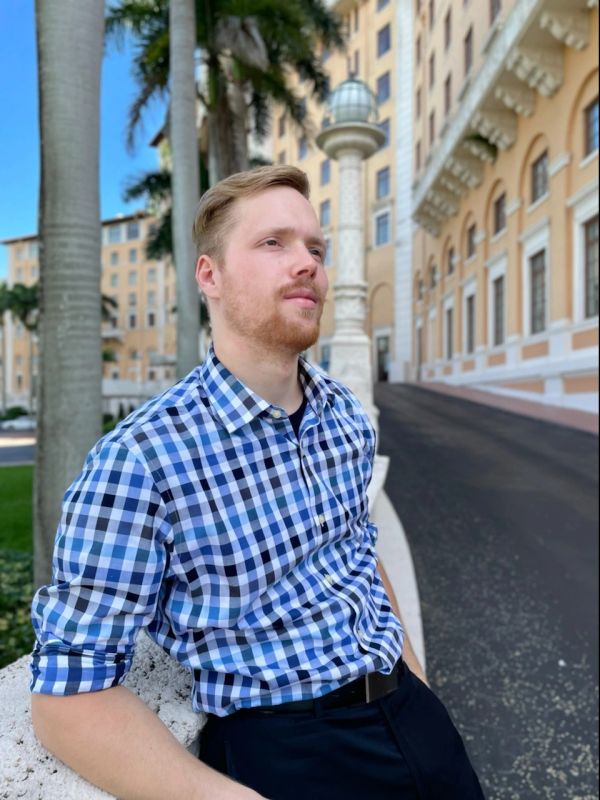 Man in a blue checkered shirt leaning against a stone ledge outdoors.