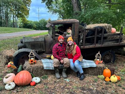 Couple posing with pumpkins and hayride truck in fall setting.