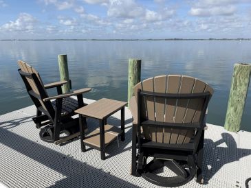 Two chairs and a table on a dock by calm water under a blue sky.