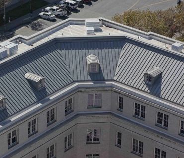 Aerial view of a building's metal roof with dormer windows and parking area.