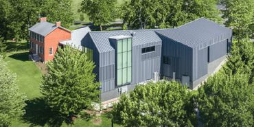 A modern gray building next to a traditional red brick house surrounded by trees.