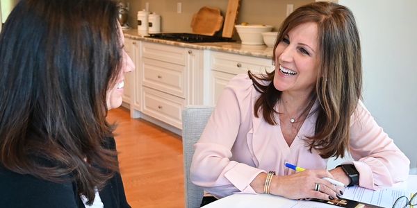 Two women smiling and conversing at a kitchen table with documents.