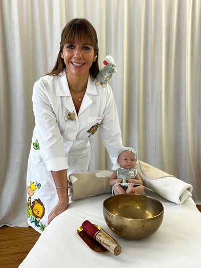 Smiling woman in a decorated white coat with a baby doll and a brass singing bowl.