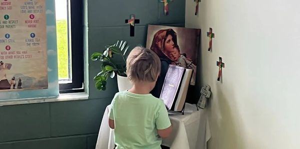 A child stands facing a small religious altar with a Bible and a painting of Mary and baby Jesus.