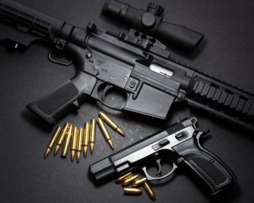A rifle and a pistol with bullets on a dark surface.