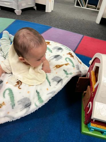 Baby lying on a colorful play mat, looking at a toy barn.