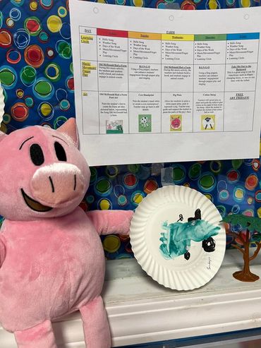 A plush pig holding a paper plate with a child's painted farm animal art.