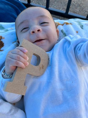 Smiling baby holding a wooden letter P while lying on a soft blanket.
