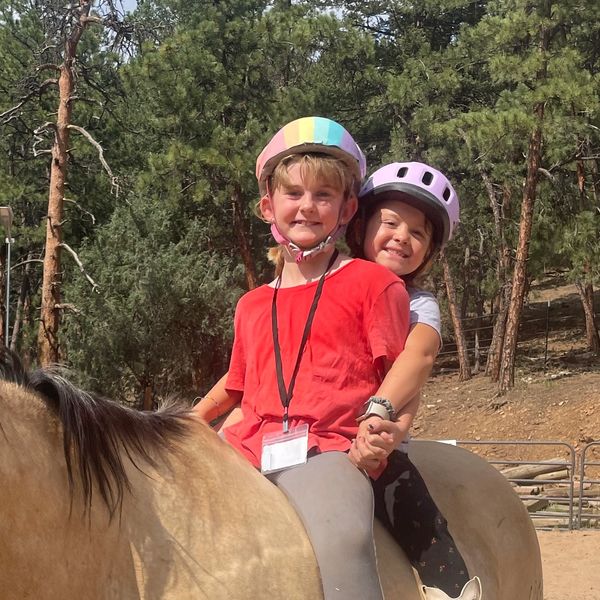 Two children riding a horse with helmets in a sandy arena.