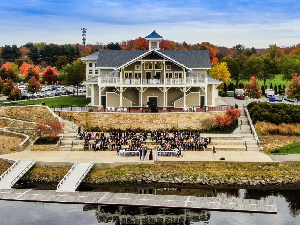 Outdoor gathering in front of a large building by the water with autumn foliage.