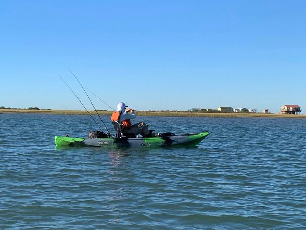 Person fishing on a green kayak in calm water under a clear blue sky.