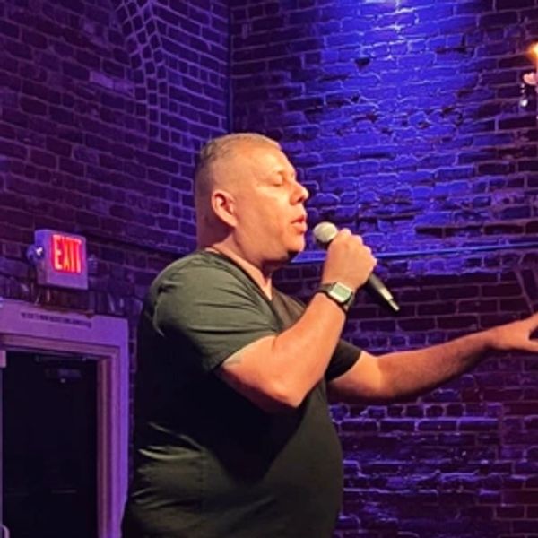 Man singing into a microphone against a brick wall backdrop.