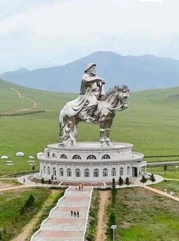 Giant equestrian statue on a circular building in a vast green landscape.
