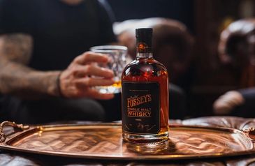 A bottle of Fossey's single malt whisky on a tray with a person holding a glass in the background.