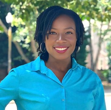 Smiling woman in a bright blue shirt outdoors with greenery in the background.