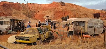 Film crew working on a desert set with old cars and trailers.