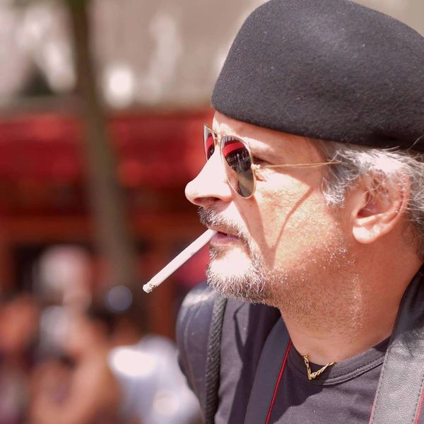 Man in sunglasses and beret smoking a cigarette outdoors.