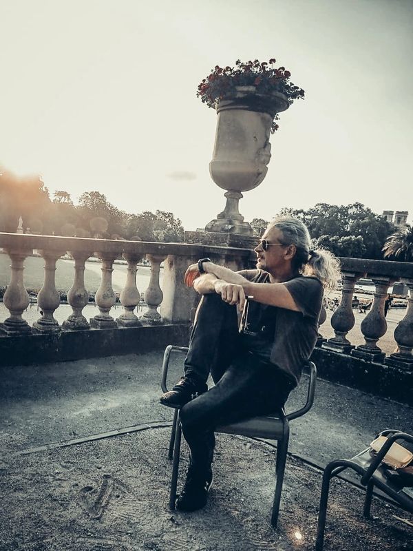 A man with long hair and sunglasses sits thoughtfully on a chair in a sunlit outdoor setting.