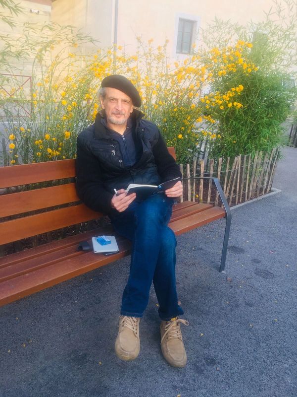 Man in a black beret sitting on a bench with a notebook, surrounded by yellow flowers.