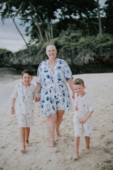 A smiling woman walks barefoot on the beach, holding hands with two young boys.
