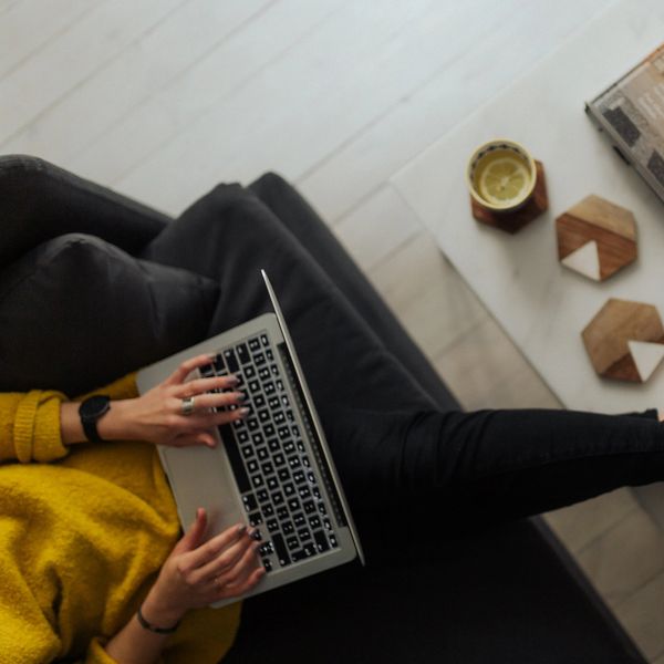 Person in black sneakers and yellow sweater works on laptop on couch.