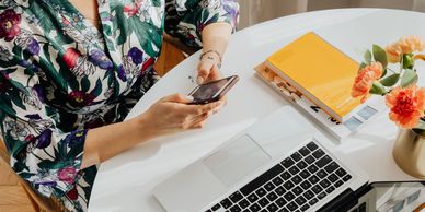 Person using smartphone at a white table with laptop and flowers.