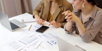 Two women working on financial documents with laptops and a calculator.