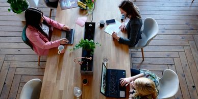 Three women working together at a wooden desk with laptops and papers.