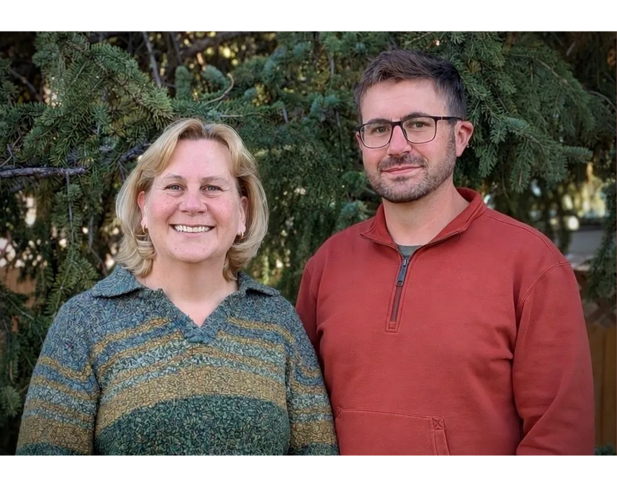 Two people smiling outdoors near pine trees.