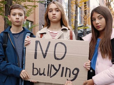 Four serious kids hold a cardboard sign reading 'NO bullying' in an outdoor urban setting.