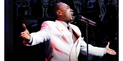 A man passionately singing on stage in a white suit.