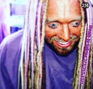 Man with star tattoos under eyes and colorful dreadlocks smiles looking down.