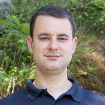 A man in a navy blue polo shirt outdoors with greenery in the background.