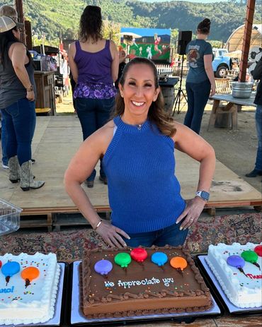 Woman smiling behind a table with decorated cakes at an outdoor event.